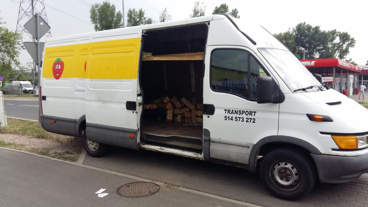 Biały samochód dostawczy z otwartymi drzwiami bocznymi, wewnątrz widoczne drewniane elementy, na boku pojazdu żółty pas z naklejką truskawki i numer telefonu do transportu.