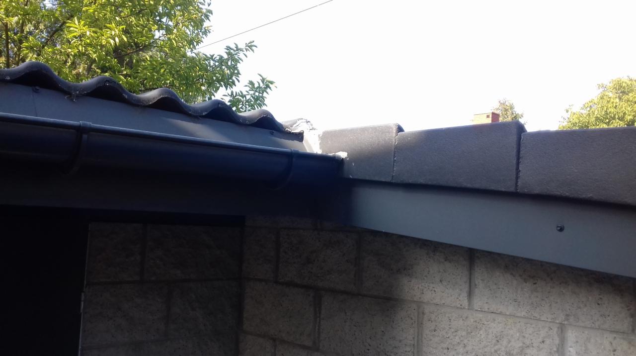 Fragment elewacji budynku z widocznym dachem pokrytym falistą dachówką, rynną oraz fragmentem ściany z szarej cegły. Widoczne uszkodzenie na styku dachu i ściany.