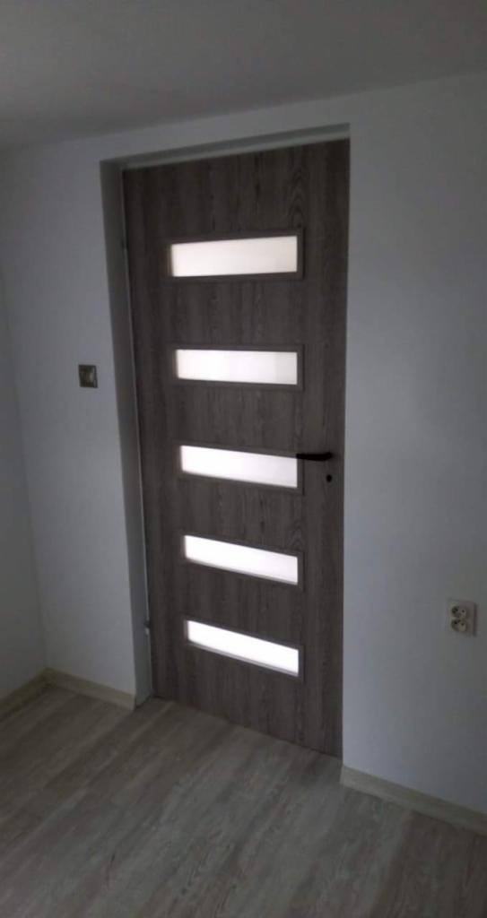 Wnętrze pokoju z zamontowanymi szarymi drzwiami z czterema poziomymi przeszkleniami, widok na podłogę wyłożoną panelami w odcieniach szarości, na ścianie widoczne gniazdko elektryczne.