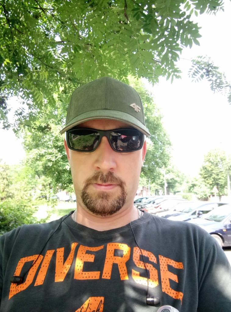 Mężczyzna w okularach przeciwsłonecznych i czapce z daszkiem, na tle zieleni drzew i parkingu. Ujęcie z dołu, widoczny fragment koszulki z napisem.