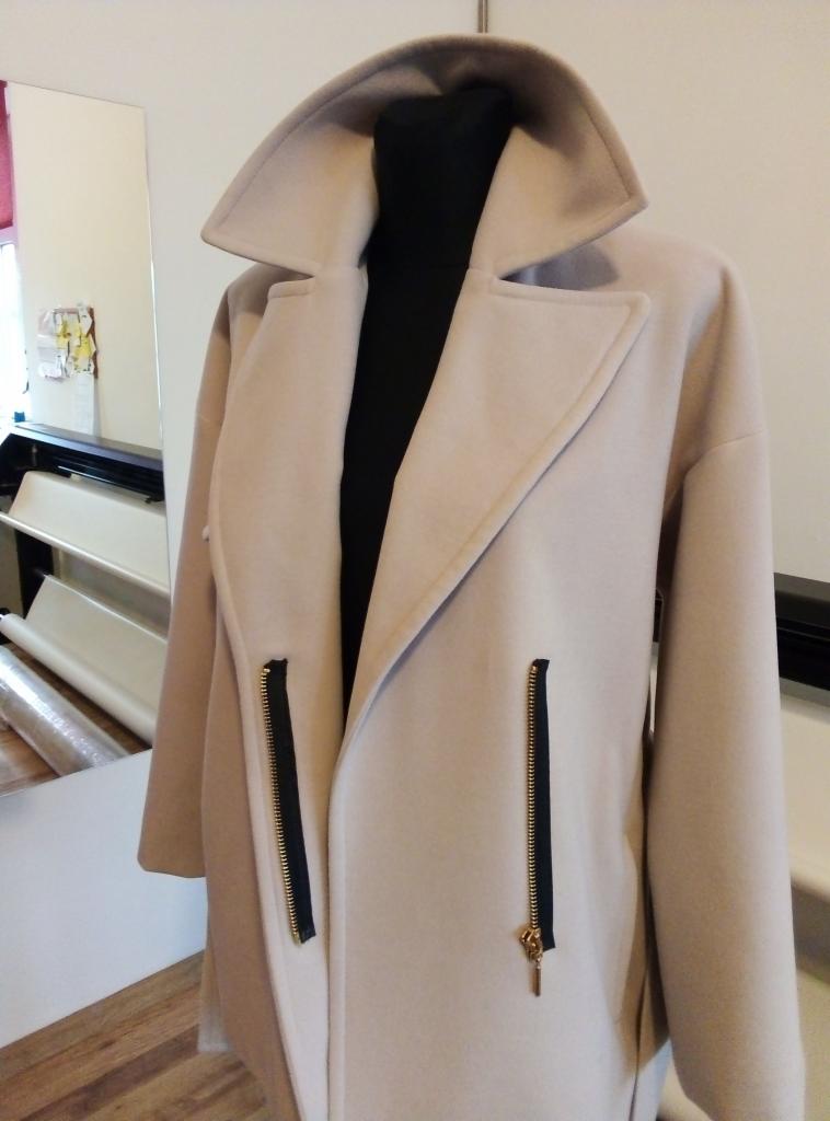 Kremowy płaszcz damski z szerokim kołnierzem i ozdobnymi, pionowymi zamkami błyskawicznymi, prezentowany na manekinie w pracowni.