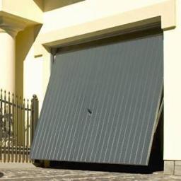 Euro Płot - Częściowo otwarta, szara brama garażowa segmentowa z pionowymi przetłoczeniami, widoczna z niskiej perspektywy, z fragmentem ogrodzenia i elewacji budynku w tle.