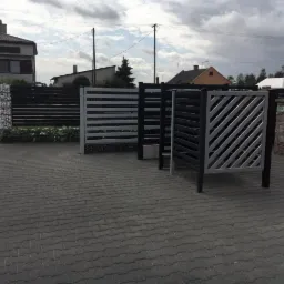 Wystawka różnych wzorów nowoczesnych ogrodzeń metalowych w kolorze czarnym i szarym, prezentowanych na szarym brukowanym podjeździe przed domem.