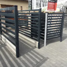 Nowoczesne, poziome przęsła ogrodzeniowe z metalu w kolorze grafitowym, zamontowane na betonowej podmurówce, z widocznym fragmentem reklamy producenta w tle.