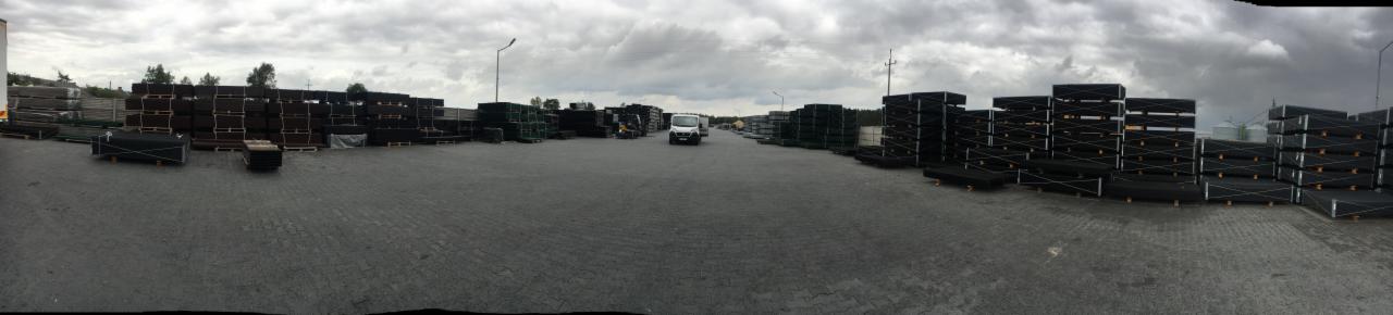 Szeroki kadr na plac składowy z ułożonymi w stosy panelami ogrodzeniowymi w czarnym kolorze, z białym samochodem dostawczym pośrodku i pochmurnym niebem w tle.