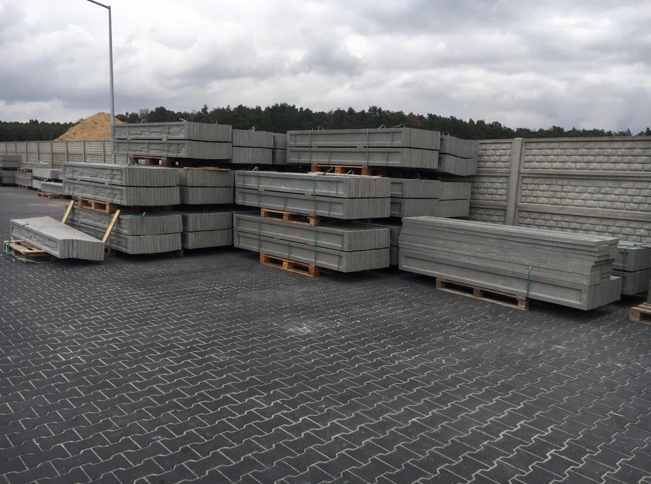 Składowane betonowe prefabrykaty ogrodzeniowe na paletach, widoczne elementy gotowego ogrodzenia z fakturą cegły, w tle hałda piasku i las.