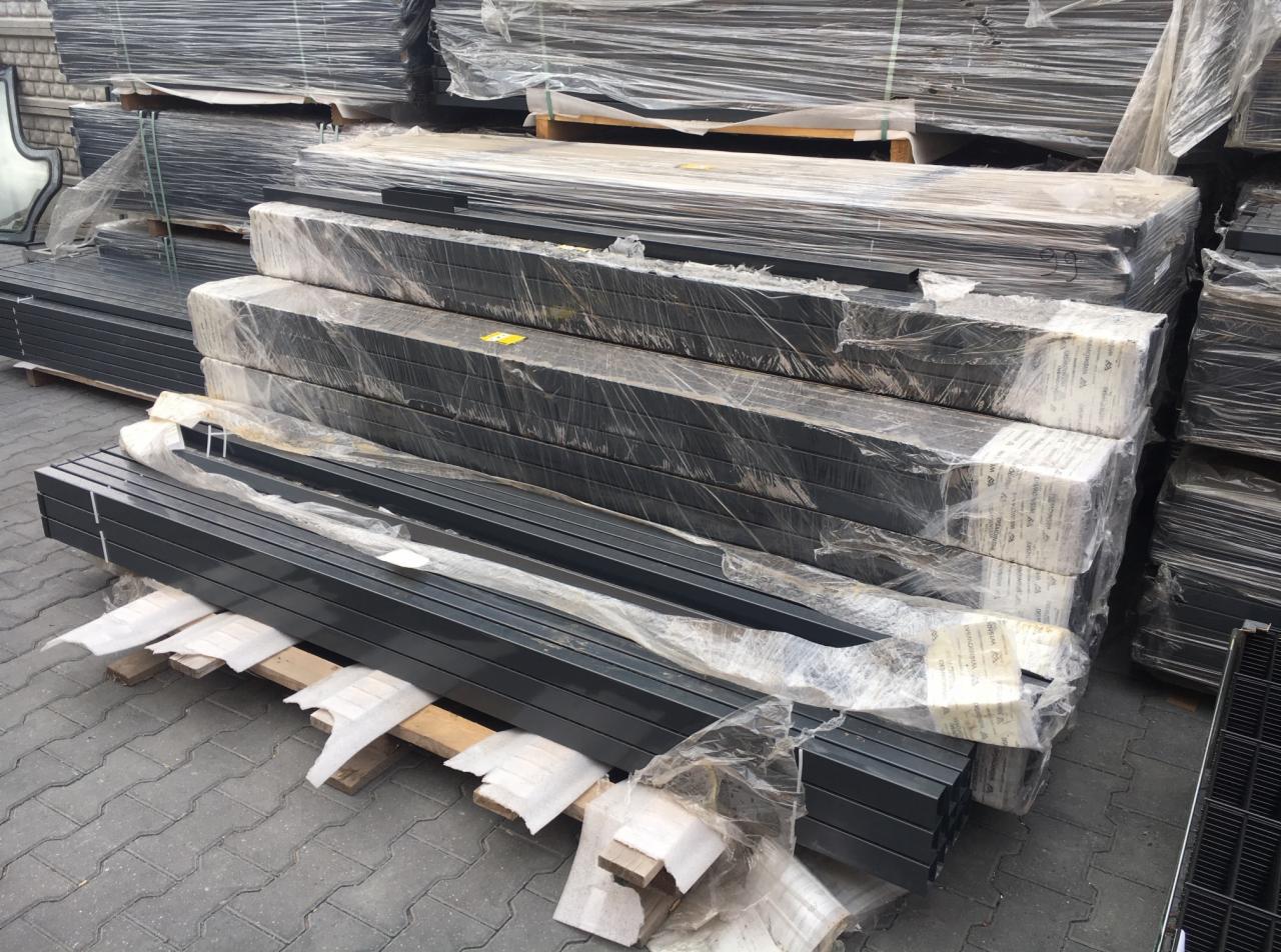 Stosy zabezpieczonych folią i taśmą pakietów metalowych elementów ogrodzeniowych lub bramowych, ułożonych na paletach na zewnątrz.