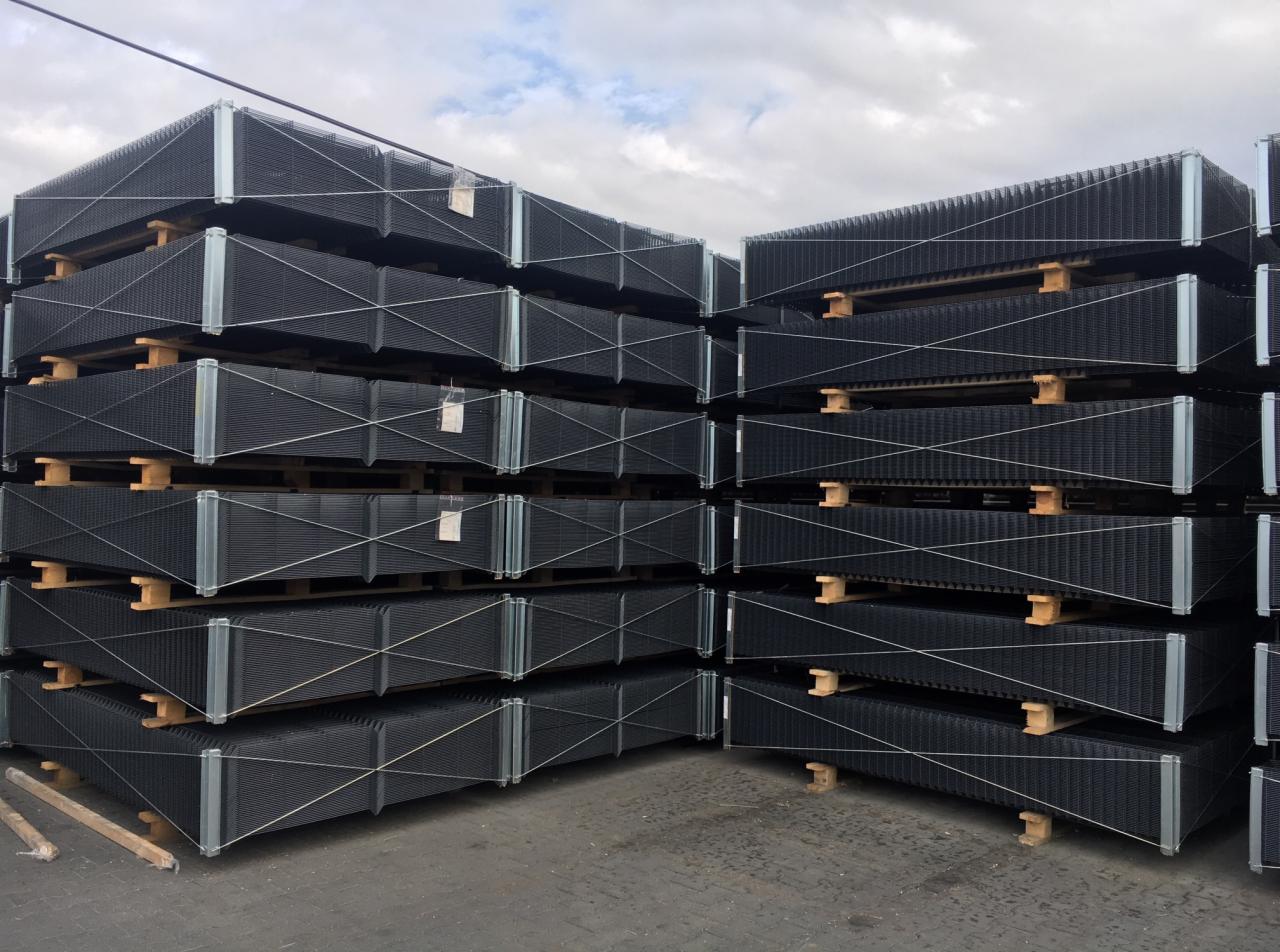 Sztaple czarnych paneli ogrodzeniowych z metalową ramą, ułożone na drewnianych paletach, składowane na zewnątrz pod pochmurnym niebem.