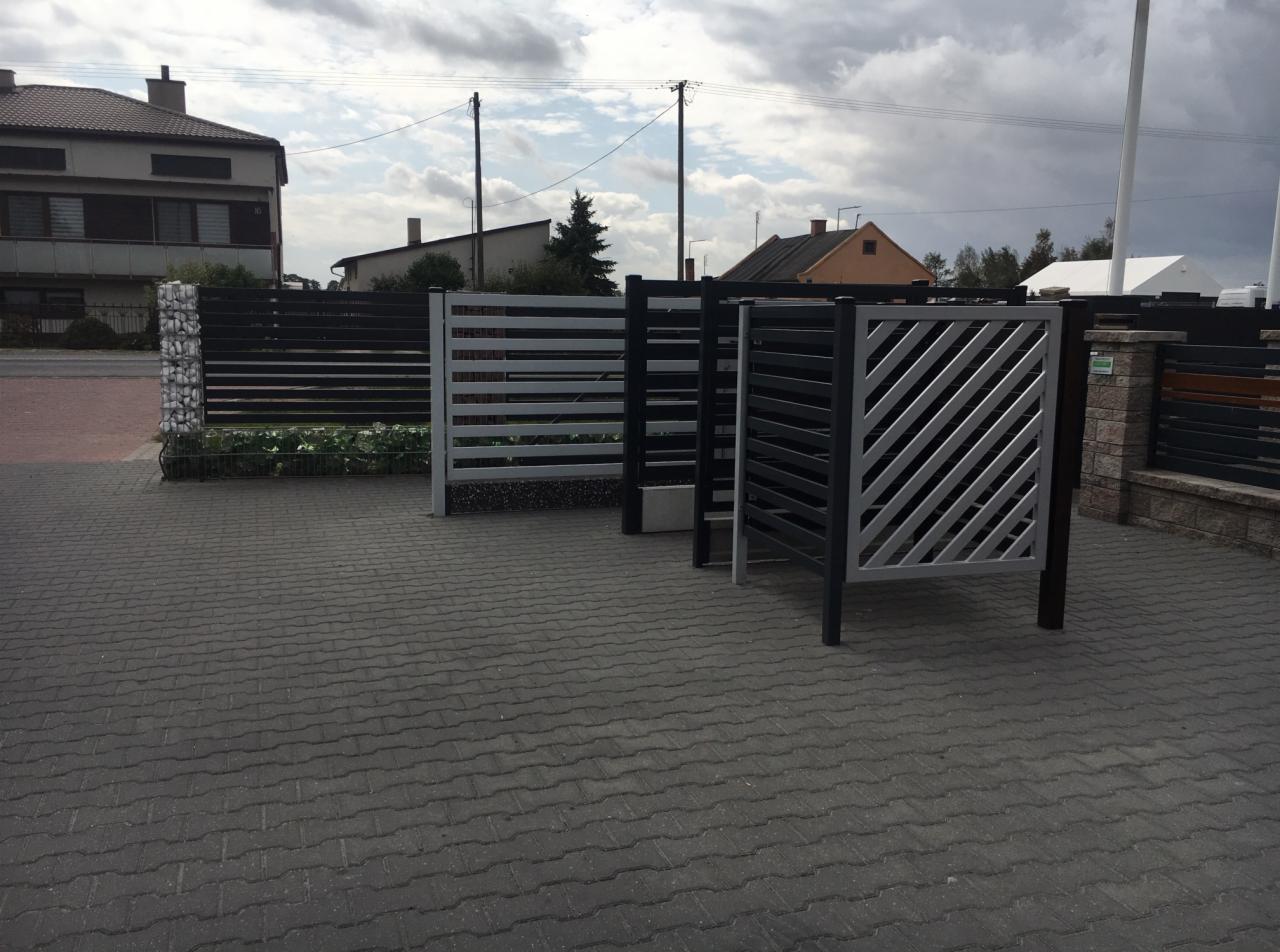 Wystawka różnych wzorów nowoczesnych ogrodzeń metalowych w kolorze czarnym i szarym, prezentowanych na szarym brukowanym podjeździe przed domem.