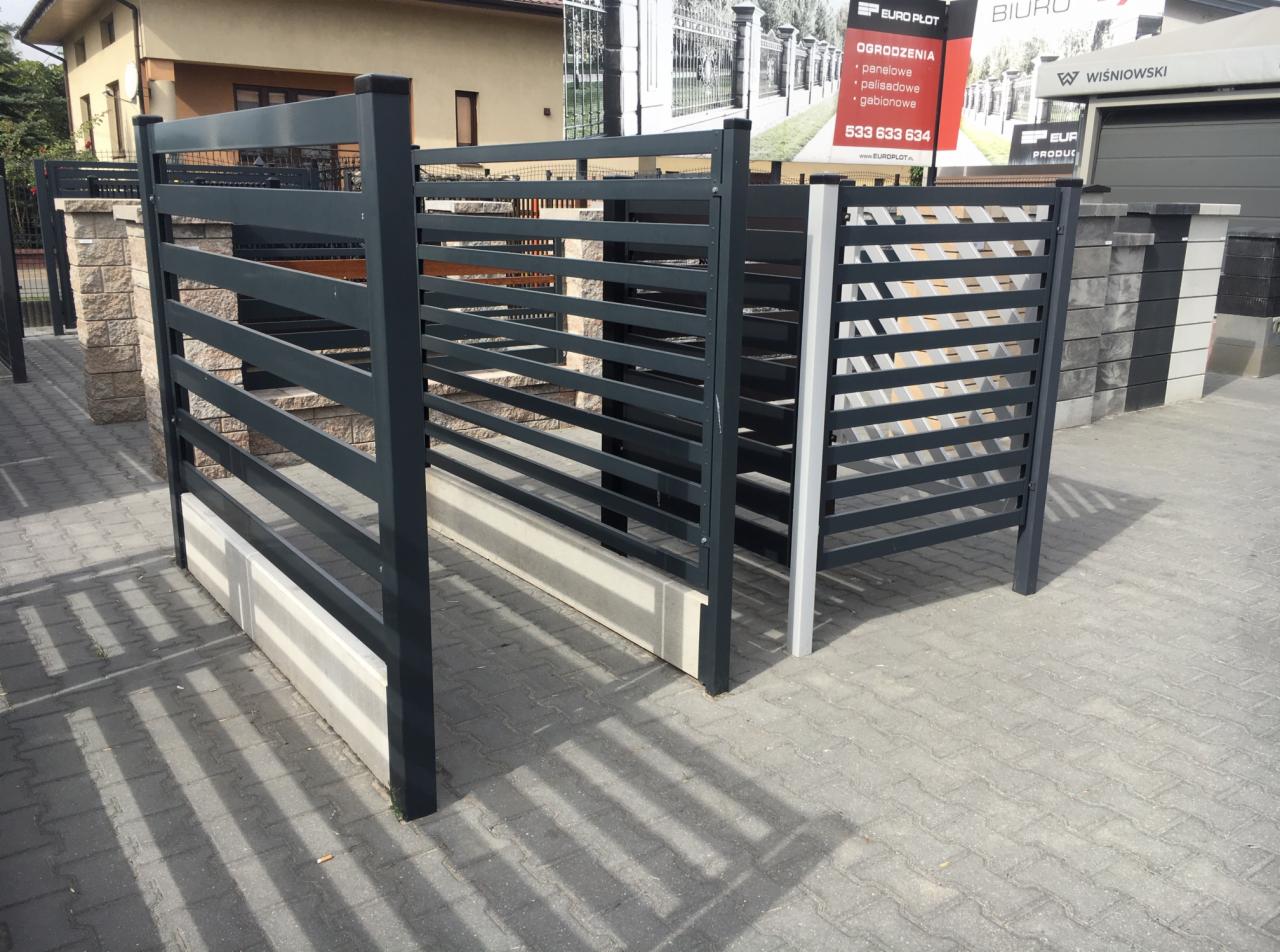 Nowoczesne, poziome przęsła ogrodzeniowe z metalu w kolorze grafitowym, zamontowane na betonowej podmurówce, z widocznym fragmentem reklamy producenta w tle.