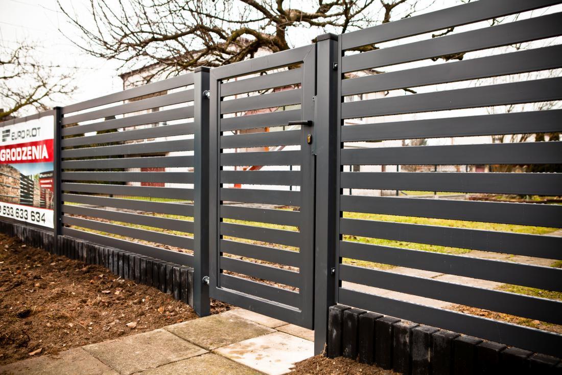Nowoczesne ogrodzenie panelowe z poziomych, szarych profili z furtką i tablicą reklamową producenta po lewej stronie. Widoczne kostki brukowe i fragment trawnika.