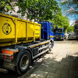 Żółty kontener wypełniony gruzem na niebieskiej ciężarówce z logo firmy 'Papaj Wynajem Kontenerów', zaparkowanej na chodniku w słoneczny dzień; w tle widoczna druga ciężarówka i budynek.