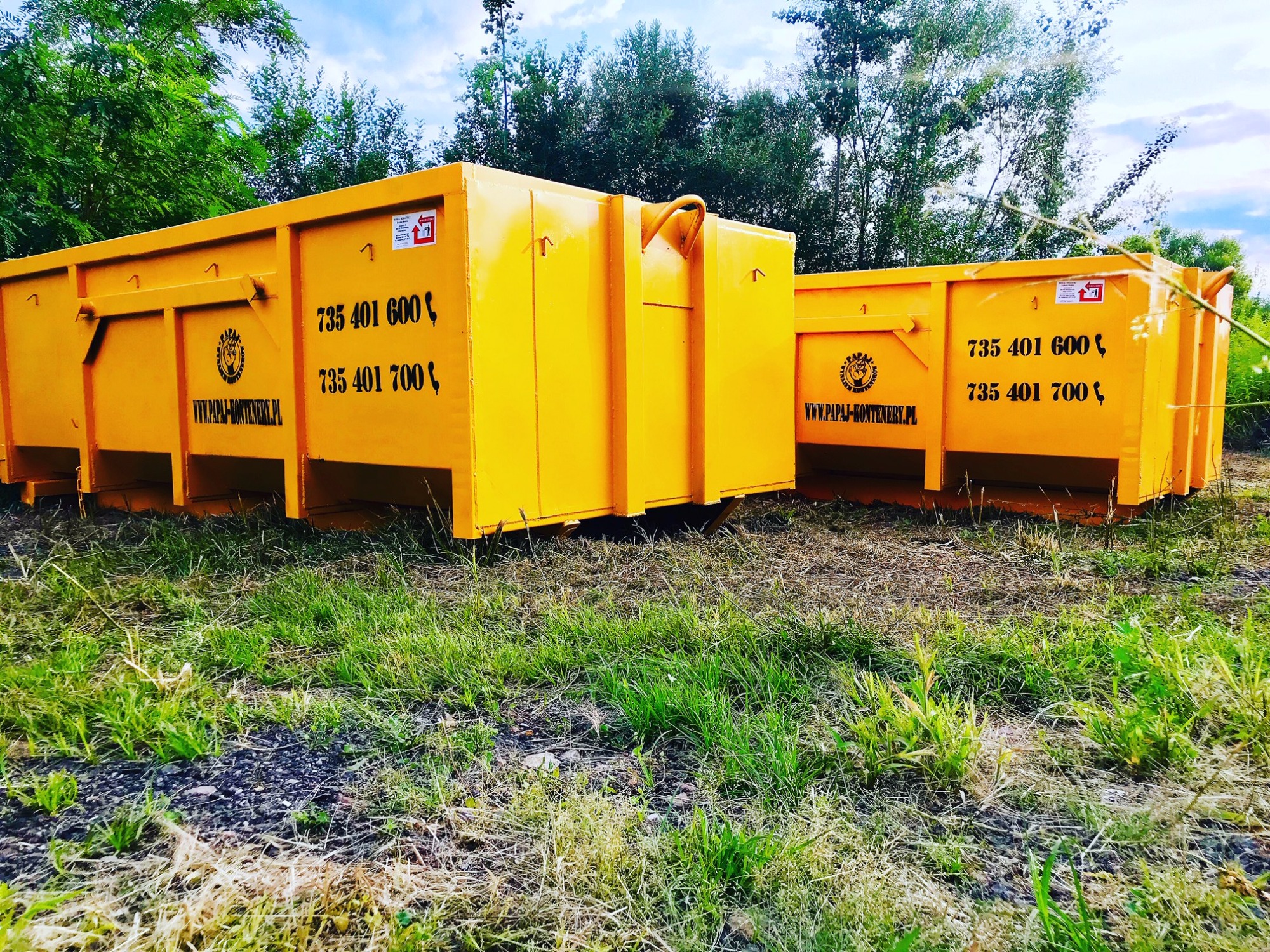 Dwa żółte, puste kontenery na gruz ustawione na trawiastym terenie, z widocznym numerem telefonu i adresem strony internetowej firmy.