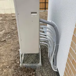 Zewnętrzna jednostka pompy ciepła zamontowana na metalowej konstrukcji, z licznymi przewodami w szarych peszlach, przymocowanymi do ściany budynku. Teren wokół jednostki jest częściowo utwardzony...