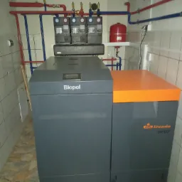 Szary piec na paliwo stałe Biopel z pomarańczową górną częścią, widoczne rury instalacji grzewczej na ścianie w pomieszczeniu z wykafelkowaną podłogą i ścianami.