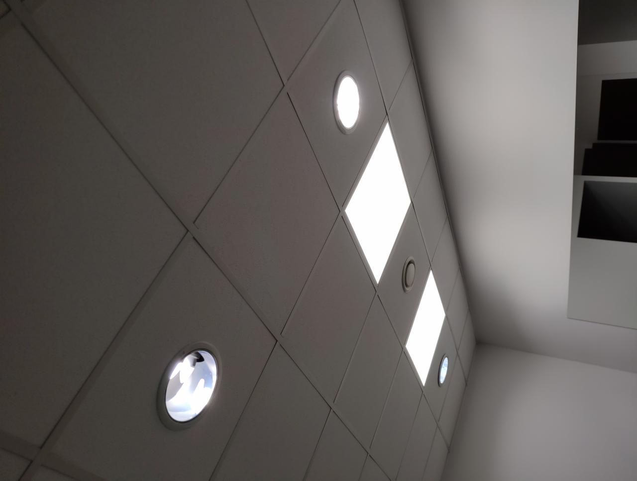 Rzut z dołu na sufit podwieszany z kwadratowymi panelami i okrągłymi lampami LED, oświetlenie włączone, widoczny fragment ściany.