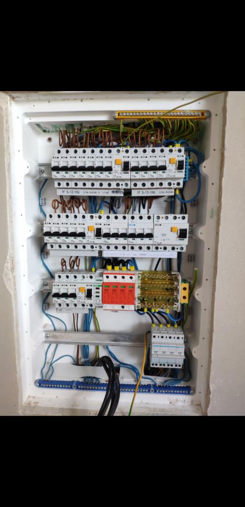 Rozdzielnica elektryczna w białej obudowie, widoczne liczne bezpieczniki, przewody w różnych kolorach, listwy zaciskowe, zabezpieczenie przeciwprzepięciowe w kolorze czerwonym, zamontowana...