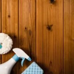 Ściereczka z mikrofibry, miotełka do kurzu, gąbka i butelka ze spryskiwaczem na tle drewnianych paneli.
