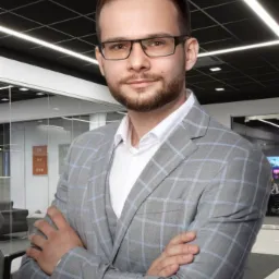 Portret mężczyzny w okularach i szarej marynarce w kratę, z rękami skrzyżowanymi na piersiach, na tle biurowego wnętrza z przeszklonymi ścianami.