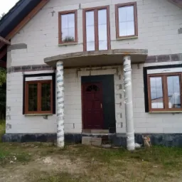 Surowy stan domu z zamontowanymi oknami z brązowymi ramami, kolumny wejściowe zabezpieczone folią ochronną, widoczny etap budowy.