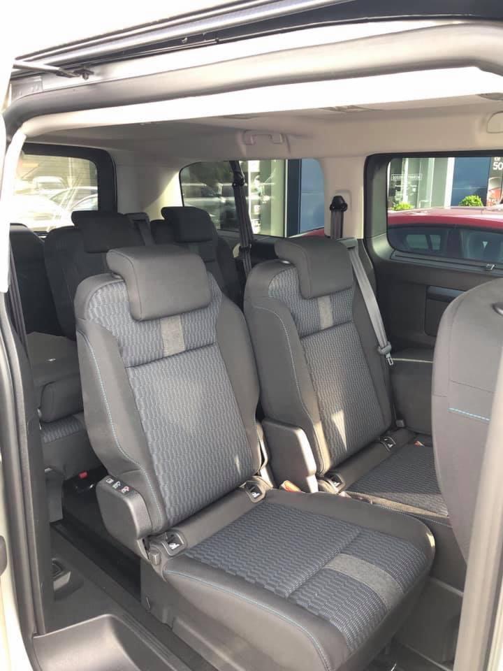 Wnętrze przestronnego busa pasażerskiego z trzema rzędami siedzeń, tapicerka w odcieniach szarości z niebieskimi przeszyciami, widok z perspektywy pasażera.