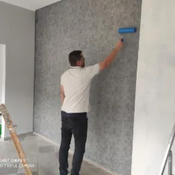 Mężczyzna maluje ścianę wałkiem na szaro, widoczna drabina malarska i puszki z farbami w tle, pomieszczenie w trakcie prac wykończeniowych.