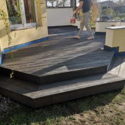 Lessandra sp. Z o.o. - Mężczyzna w jasnych spodniach i ciemnej koszulce idzie po drewnianym tarasie z puszką farby w ręku, widoczny fragment budynku z oknem i pnącą się rośliną, oraz zadaszenie tarasu.