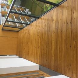 Lessandra sp. Z o.o. - Drewniana boazeria pionowa w ciepłym odcieniu brązu, z widocznymi gwoździami, tworząca ścianę balkonu z przeszklonym zadaszeniem, na tle budynku mieszkalnego.