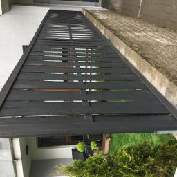 Lessandra sp. Z o.o. - Ciemnoszara drewniana konstrukcja z poziomych desek, prawdopodobnie zadaszenie lub element altany, zamontowana przy budynku z widocznym oknem, z betonową nawierzchnią obok i zielenią w tle.