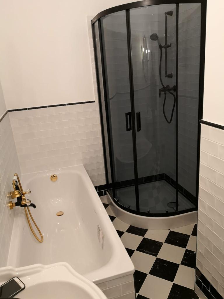 Biała wanna z złotą armaturą i czarna kabina prysznicowa w rogu łazienki z biało-czarną szachownicą na podłodze i białymi kafelkami na ścianach.