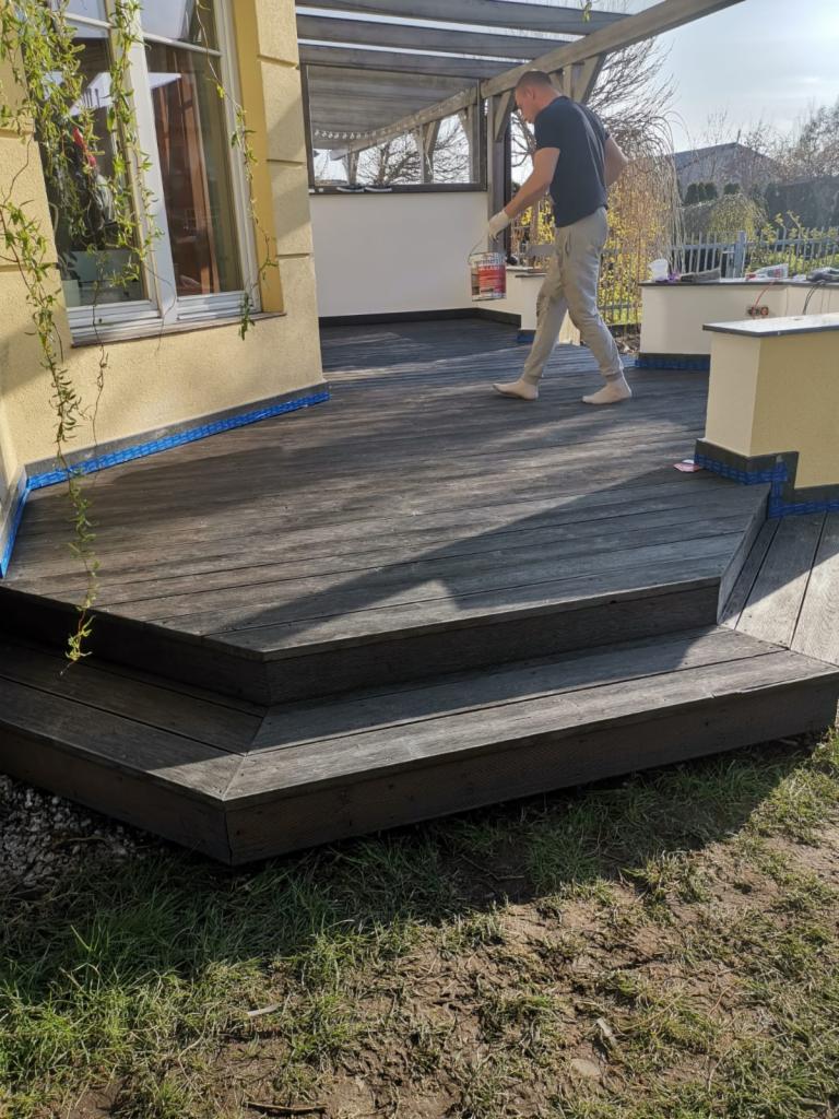 Mężczyzna w jasnych spodniach i ciemnej koszulce idzie po drewnianym tarasie z puszką farby w ręku, widoczny fragment budynku z oknem i pnącą się rośliną, oraz zadaszenie tarasu.