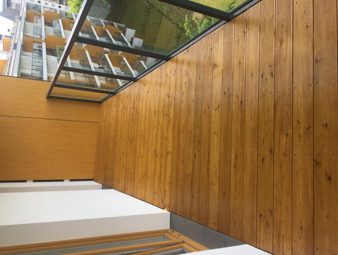 Drewniana boazeria pionowa w ciepłym odcieniu brązu, z widocznymi gwoździami, tworząca ścianę balkonu z przeszklonym zadaszeniem, na tle budynku mieszkalnego.