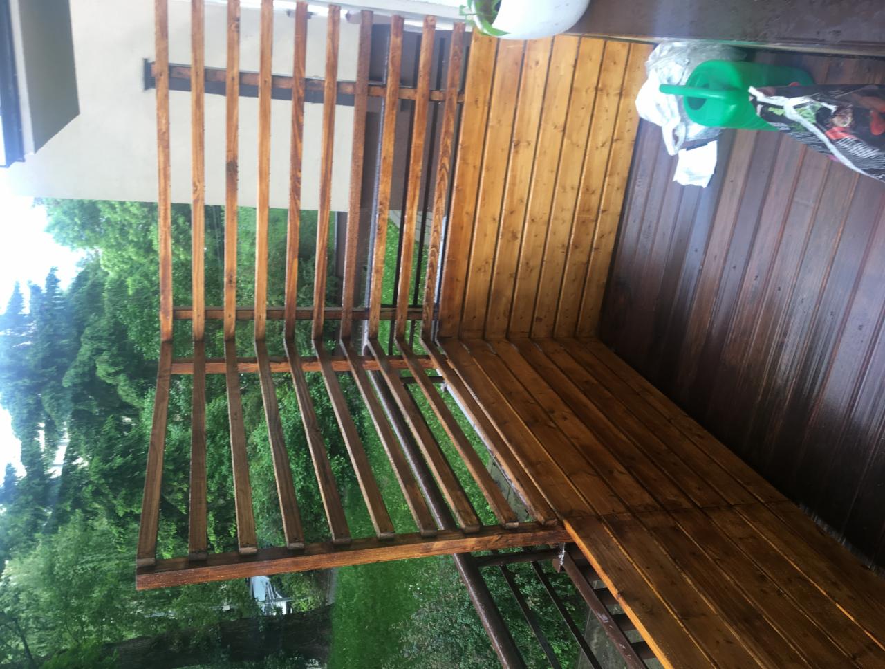 Świeżo pomalowana drewniana konstrukcja altany z widocznymi smugami mokrej farby i odbiciami, z zielonymi drzewami w tle.