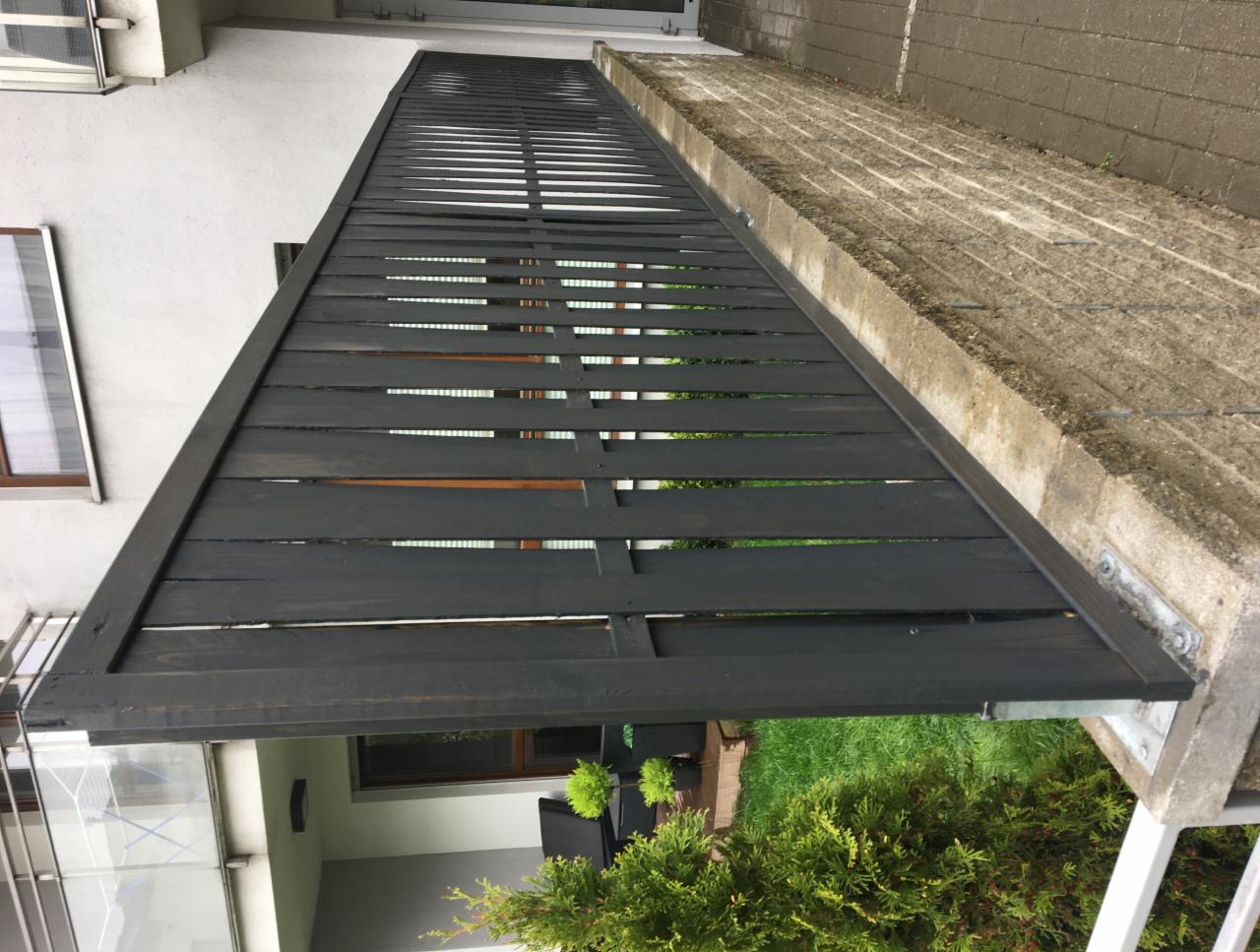 Ciemnoszara drewniana konstrukcja z poziomych desek, prawdopodobnie zadaszenie lub element altany, zamontowana przy budynku z widocznym oknem, z betonową nawierzchnią obok i zielenią w tle.