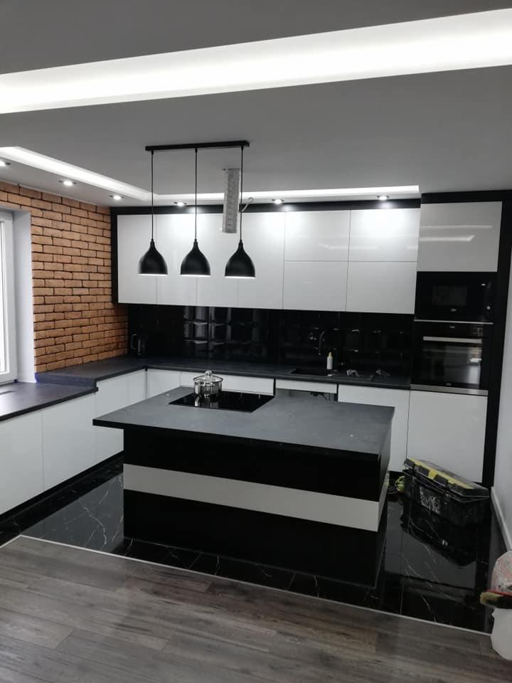 Nowoczesna kuchnia w bieli i czerni: błyszczące fronty, ceglana ściana, marmurowa podłoga i wyspa z płytą indukcyjną pod trzema wiszącymi lampami.