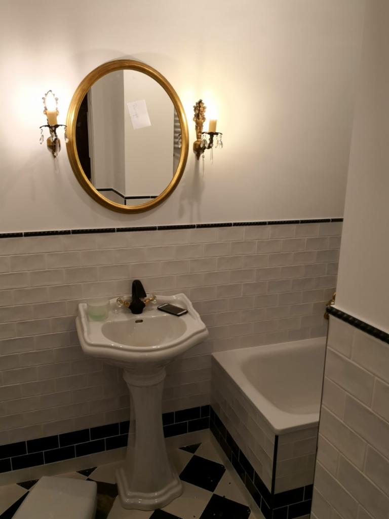 Klasyczna łazienka z czarno-białą szachownicą na podłodze, białą umywalką na postumencie, owalnym lustrem w złotej ramie i kinkietami w stylu retro.