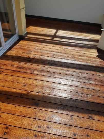 Świeżo polakierowana drewniana podłoga tarasu z widocznymi słojami drewna w ciepłym świetle słonecznym, z fragmentem okna balkonowego po lewej stronie.