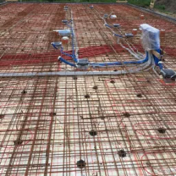 Zbrojenie płyty fundamentowej z instalacją ogrzewania podłogowego oraz wyprowadzonymi instalacjami wodno-kanalizacyjnymi, widok z góry na plac budowy.