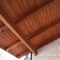 Drewniana konstrukcja dachu z widocznymi belkami nośnymi, pokryta deskami w odcieniu brązu, przylegająca do ściany z białego pustaka.