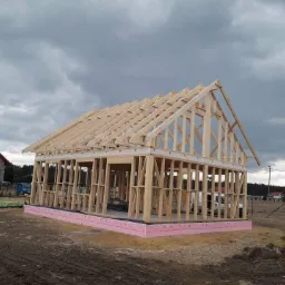 Konstrukcja drewniana domu w trakcie budowy, widoczny szkielet ścian i więźba dachowa, różowa izolacja fundamentu, pochmurne niebo w tle.