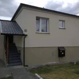 Dwukondygnacyjny dom po renowacji elewacji, z numerem 10 nad wejściem z zadaszeniem z grafitowej dachówki, jasnobeżowa fasada z ciemniejszym pasem w dolnej części, widoczna skrzynka na listy.