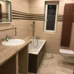 Jasna łazienka z beżowymi płytkami, brązowymi akcentami i grzejnikiem drabinkowym, widoczna wanna obudowana ciemnym drewnem oraz umywalka z blatem imitującym marmur, okno z dekoracyjną folią.