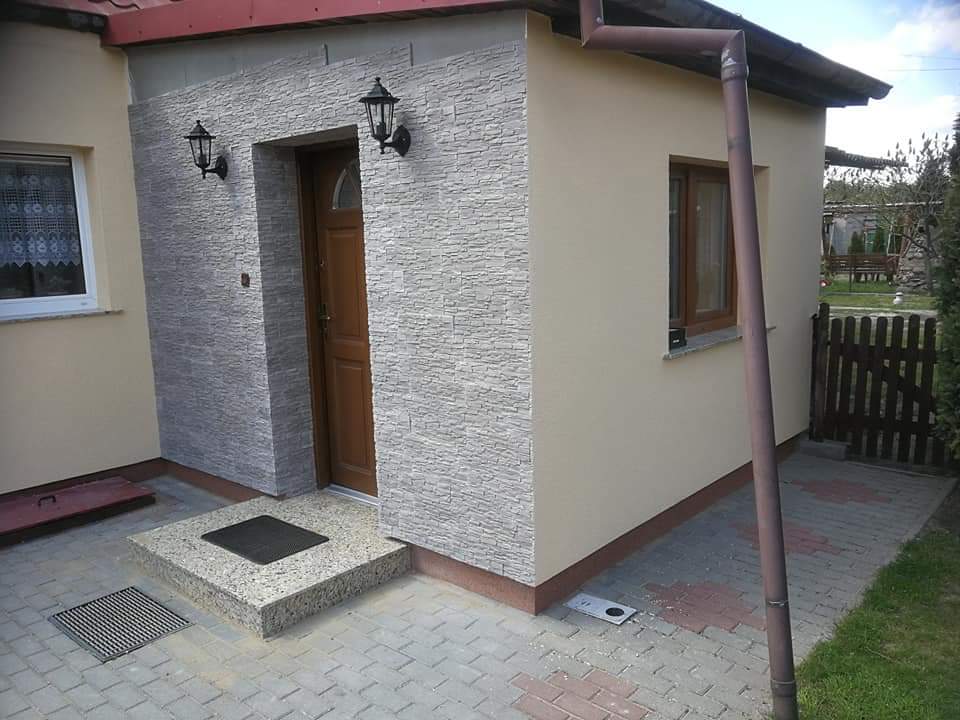 Wejście do budynku z elewacją wykończoną kamieniem dekoracyjnym, oświetlone dwoma czarnymi kinkietami, z brązowymi drzwiami i kremową ścianą boczną z brązową ramą okienną oraz rynną.