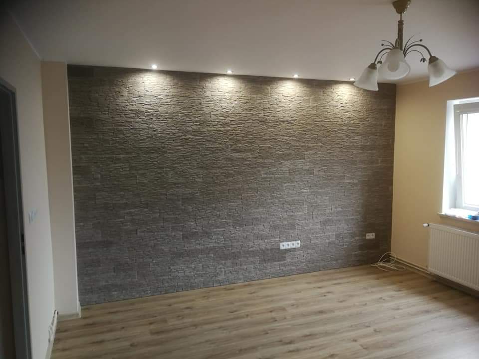 Ściana w salonie wykończona szarą okładziną imitującą kamień, oświetlona punktowymi lampami sufitowymi, z widocznymi gniazdkami elektrycznymi i drewnopodobną podłogą.