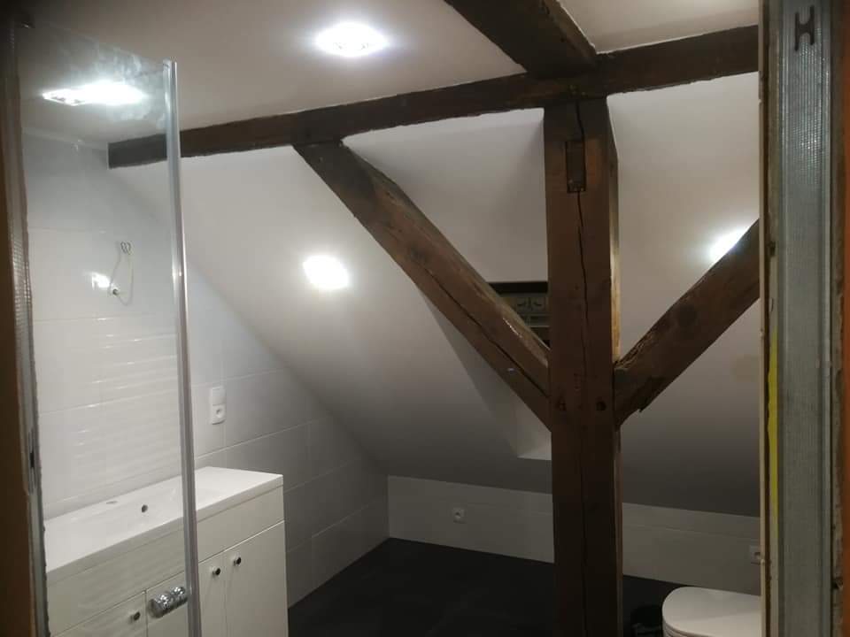 Wykończona łazienka na poddaszu z widocznymi drewnianymi belkami konstrukcyjnymi, białą armaturą i szarą podłogą, oświetlona punktowymi lampami sufitowymi.