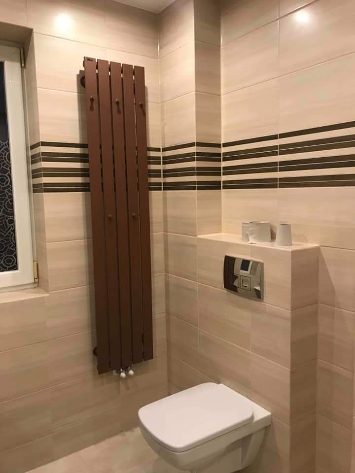 Beżowa łazienka z wiszącą toaletą, brązowym grzejnikiem drabinkowym i poziomymi paskami dekoracyjnymi na płytkach.