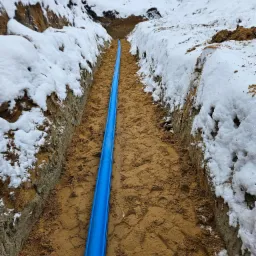 Niebieska rura w wykopie zasypanym piaskiem, obok ściany wykopu pokryte śniegiem, zimowy krajobraz.