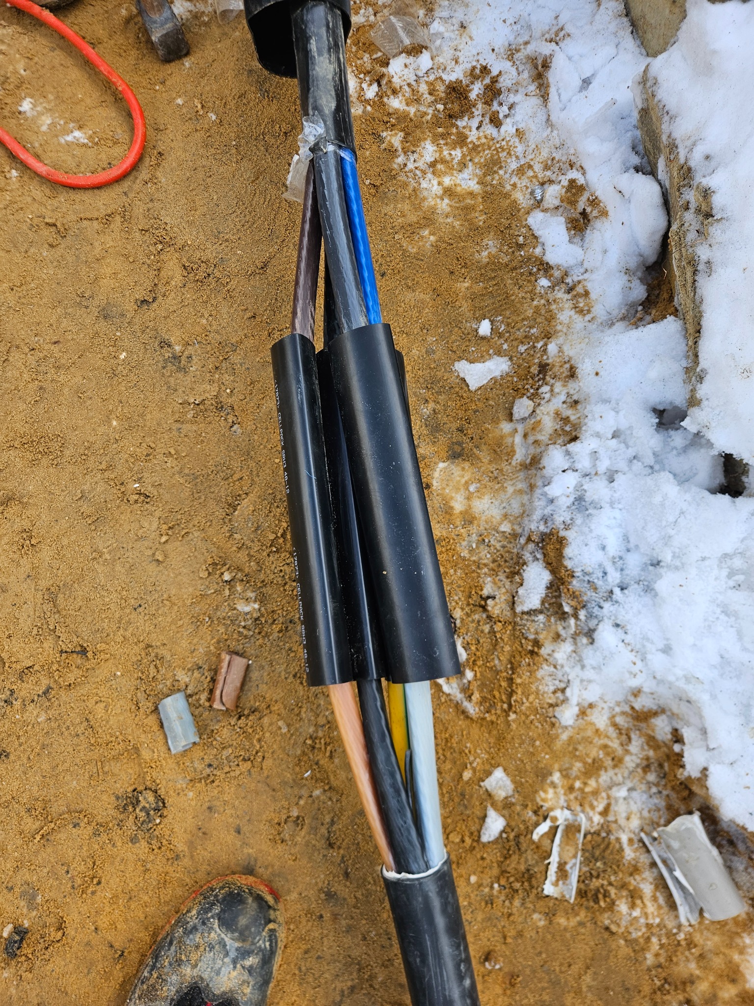 Kabel elektryczny z rozdzielonymi przewodami w czarnej osłonie, częściowo zakopany w piasku obok resztek śniegu i narzędzi, widok z góry na placu budowy.