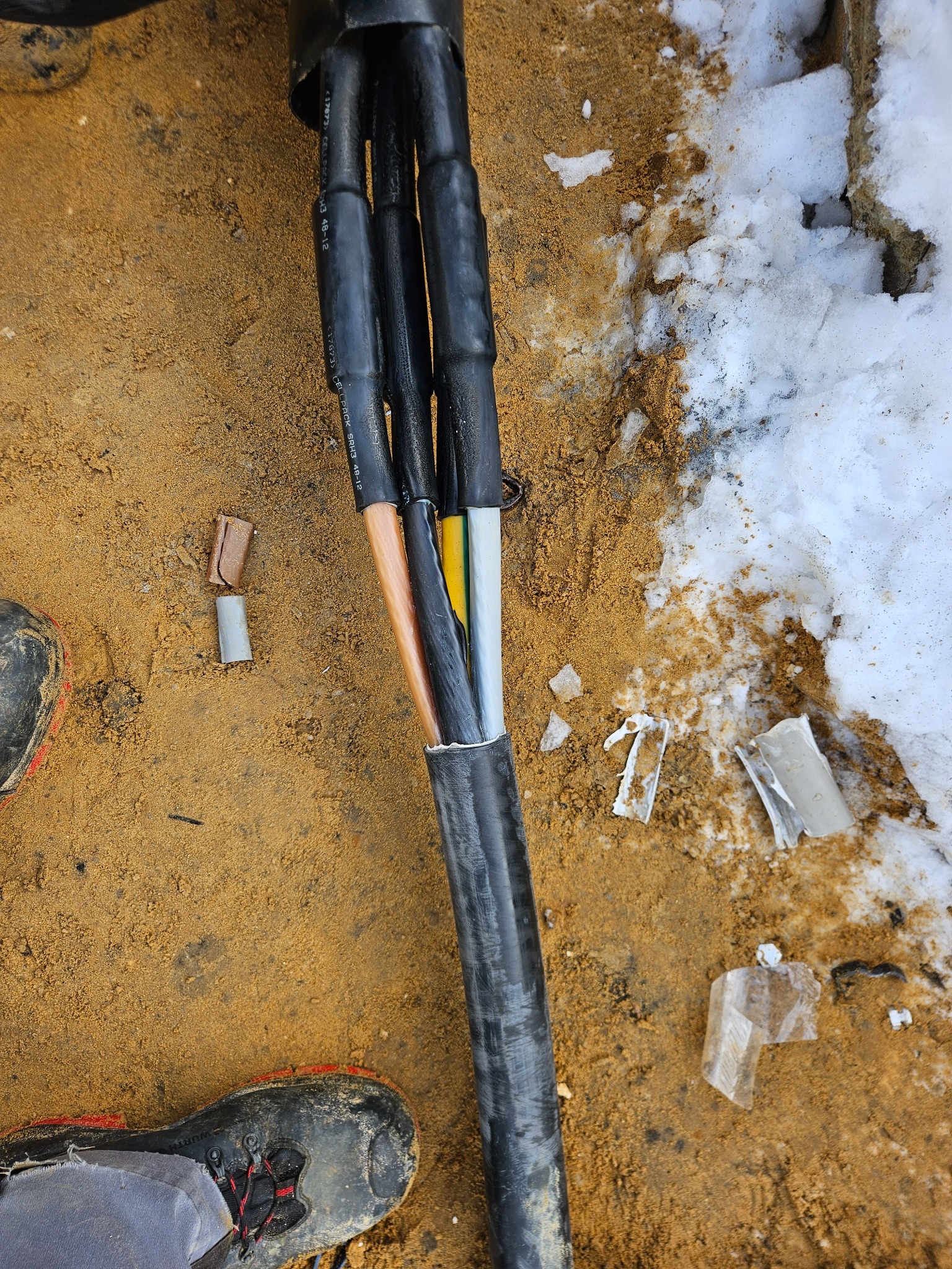 Kabel elektryczny wielożyłowy z odsłoniętymi żyłami w izolacji kolorowej (miedzianej, żółtej, szarej) leżący na ziemi z piaskiem i resztkami śniegu, obok fragment buta roboczego i odcięte końcówki