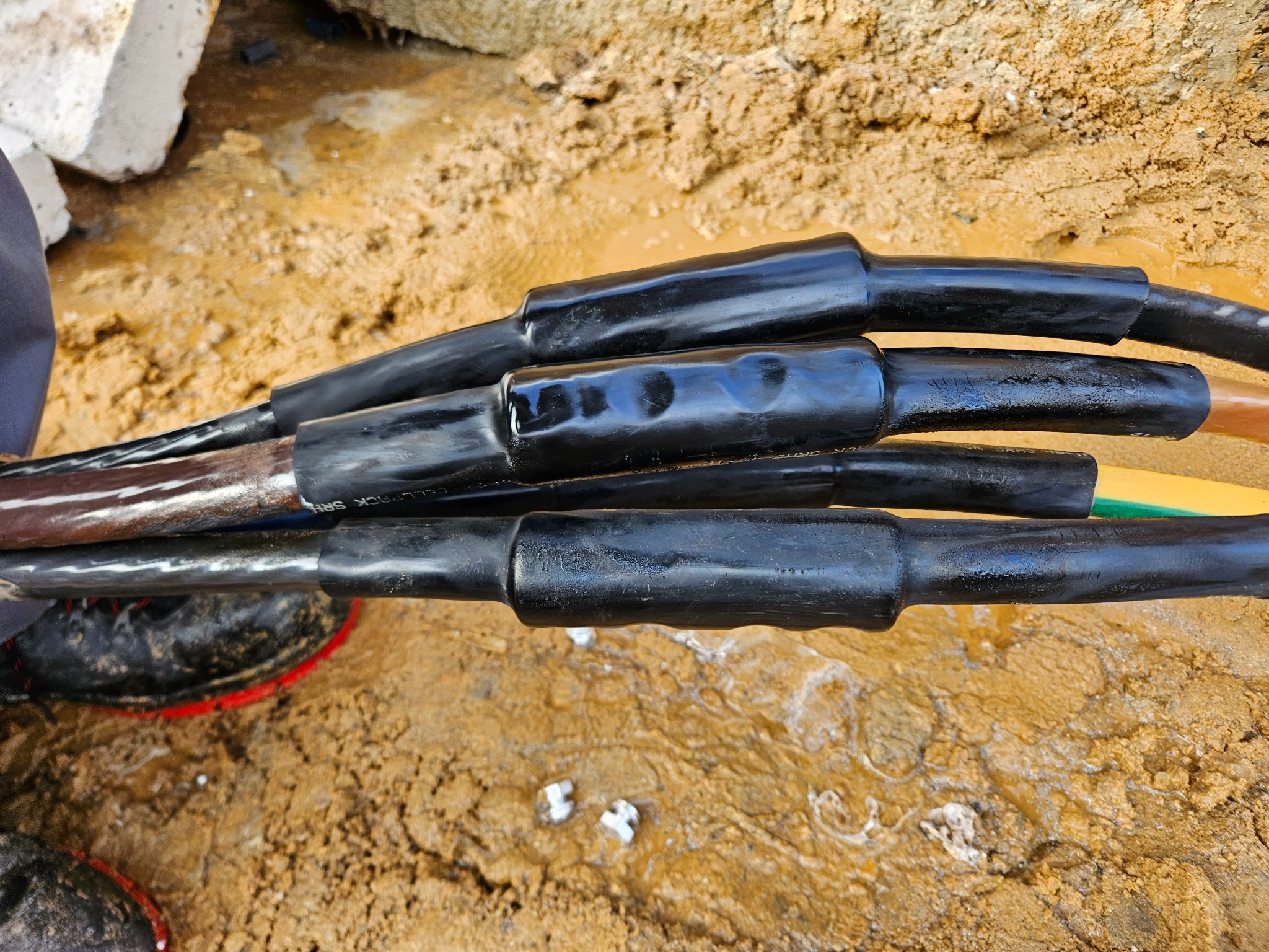Połączenie kabli elektrycznych ziemnych, z czarnymi, szczelnymi mufami termokurczliwymi, częściowo zanurzone w błotnistym wykopie, widoczne fragmenty kolorowych izolacji.